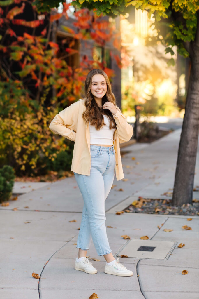 a girl in a yellow shacket walking down sidewalk with colorful leaves during golden hour senior photos 