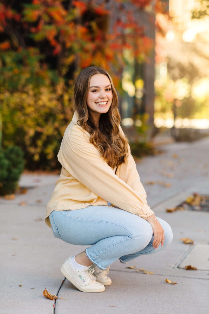a girl in a yellow shacket kneeling down on sidewalk with colorful leaves during golden hour senior photos 